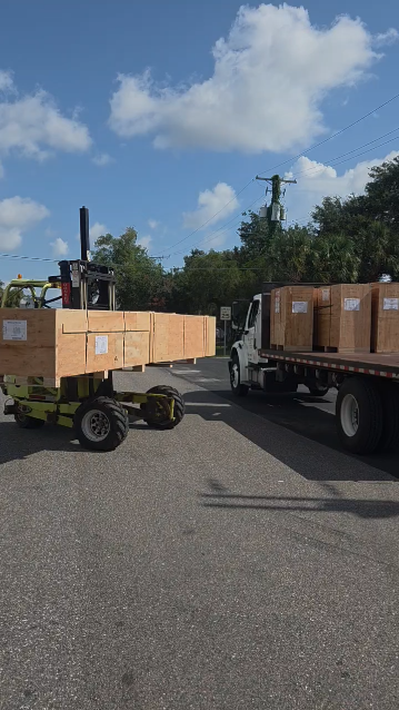 JIT truck-mounted forklift carrying a long wooden crate beside a loaded flatbed for final-mile delivery
