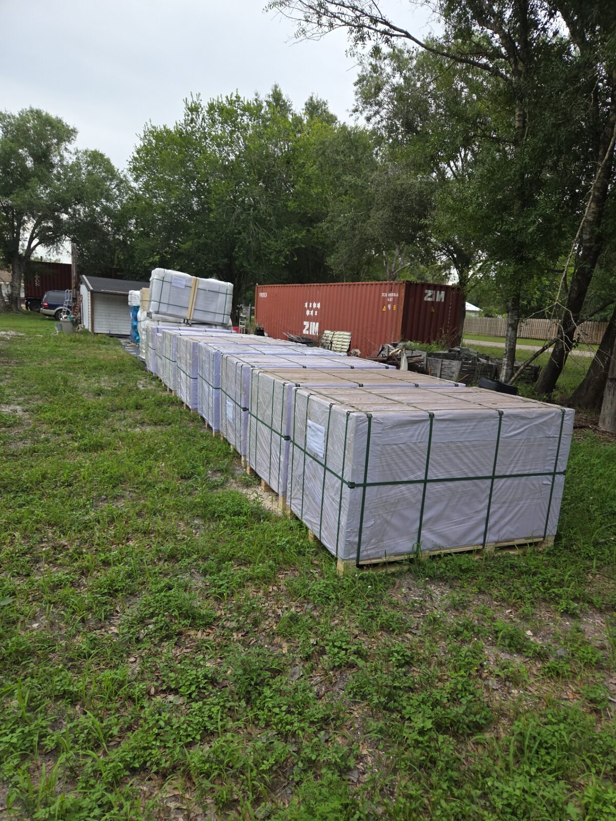 Long row of tarp-wrapped material pallets staged on a grass lot near a shipping container